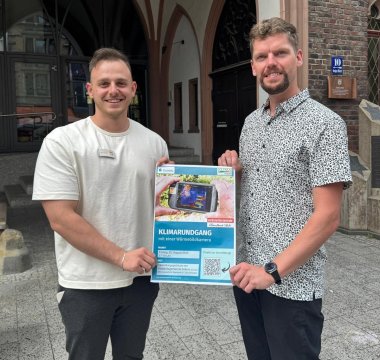 Zwei Klimaschutzmanager stehen vor dem Rathaus in Montabaur und halten die Einladung zum Klimarundgang hoch.